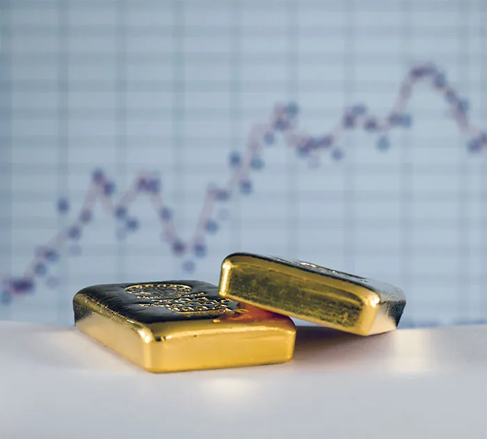 Two gold bullion bars on a desk in front of a blurred market chart